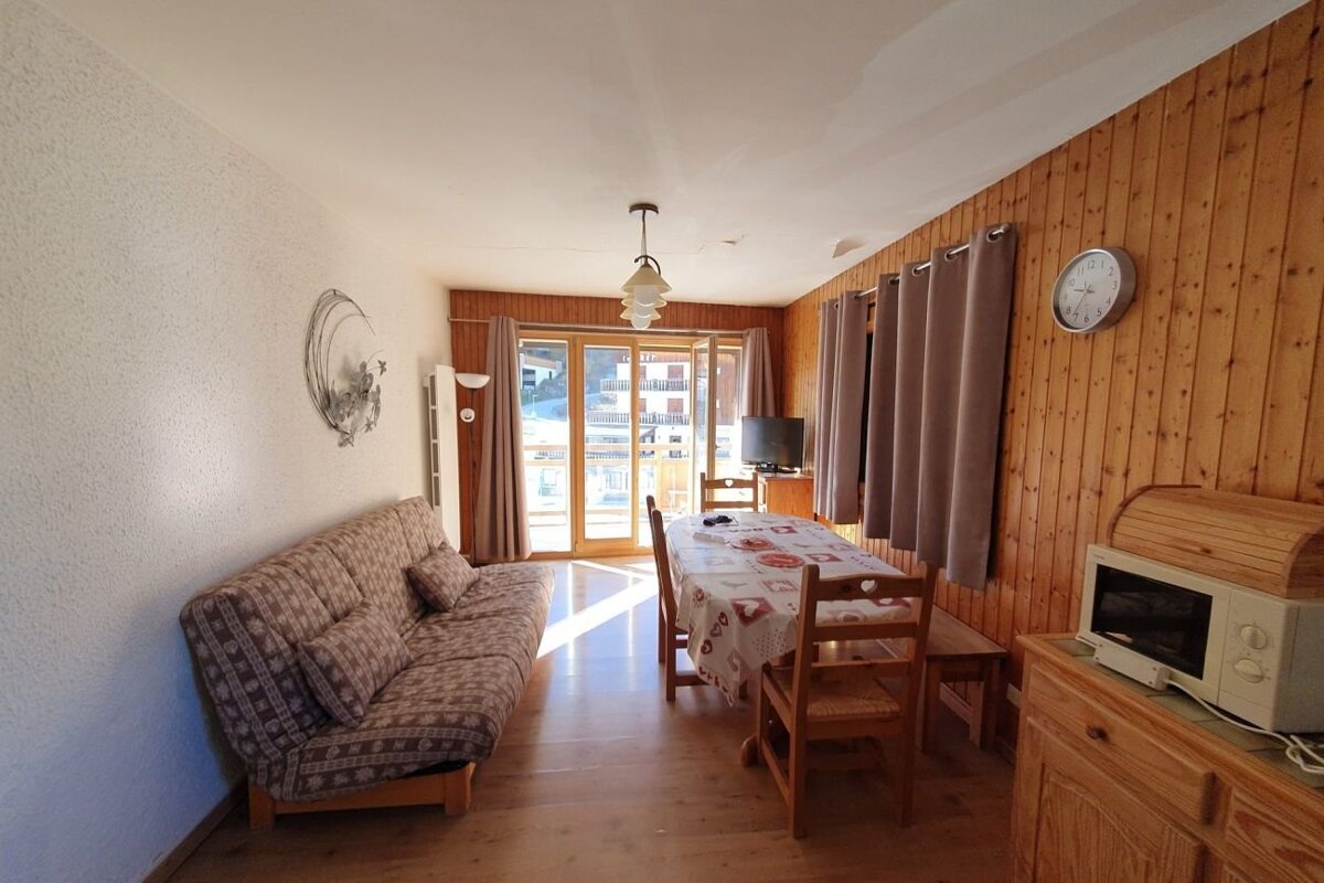 Cozy cabin-style room featuring wood-paneled walls, a patterned sofa, a dining table, and a glass balcony door letting in sunlight.