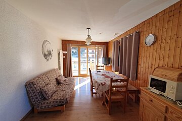 Cozy cabin-style room featuring wood-paneled walls, a patterned sofa, a dining table, and a glass balcony door letting in sunlight.