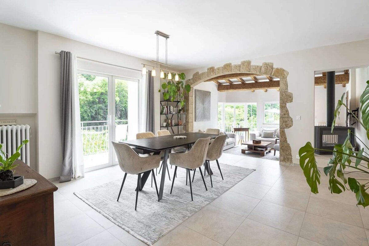 A bright, open-plan dining room with a modern table and chairs, featuring a rustic stone archway leading to a living area with large windows and lush greenery outside.