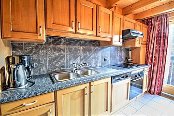 A compact kitchen with wood cabinets, dark tiled backsplash, sink, stove, oven, kettle, coffee maker, and red patterned curtains by a window.