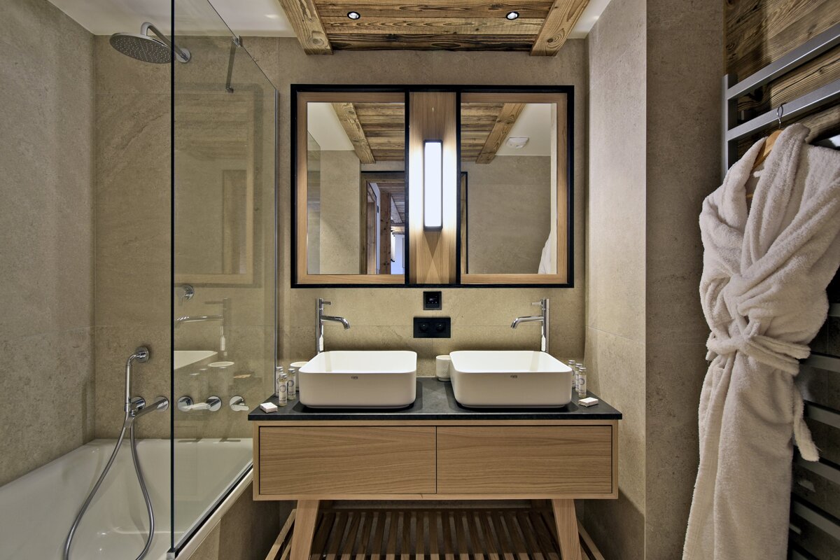 Modern bathroom with a double sink vanity, wood accents, and a glass-enclosed shower/tub. Earthy tiles and wooden beams create a warm, inviting feel.