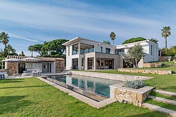 A contemporary white villa with a large rectangular pool, green lawn, and surrounding trees under a clear blue sky.