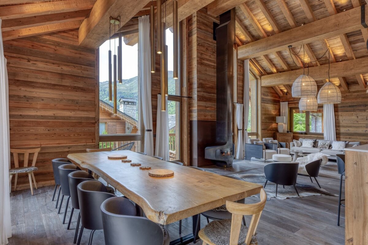 Rustic luxury chalet interior featuring a long wood dining table, suspended fireplace, comfy lounge, and mountain views.