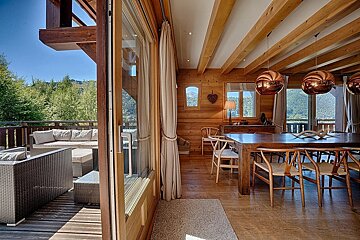A warm wooden chalet interior with a dining room and an outdoor terrace overlooking lush green mountains under a clear sky.