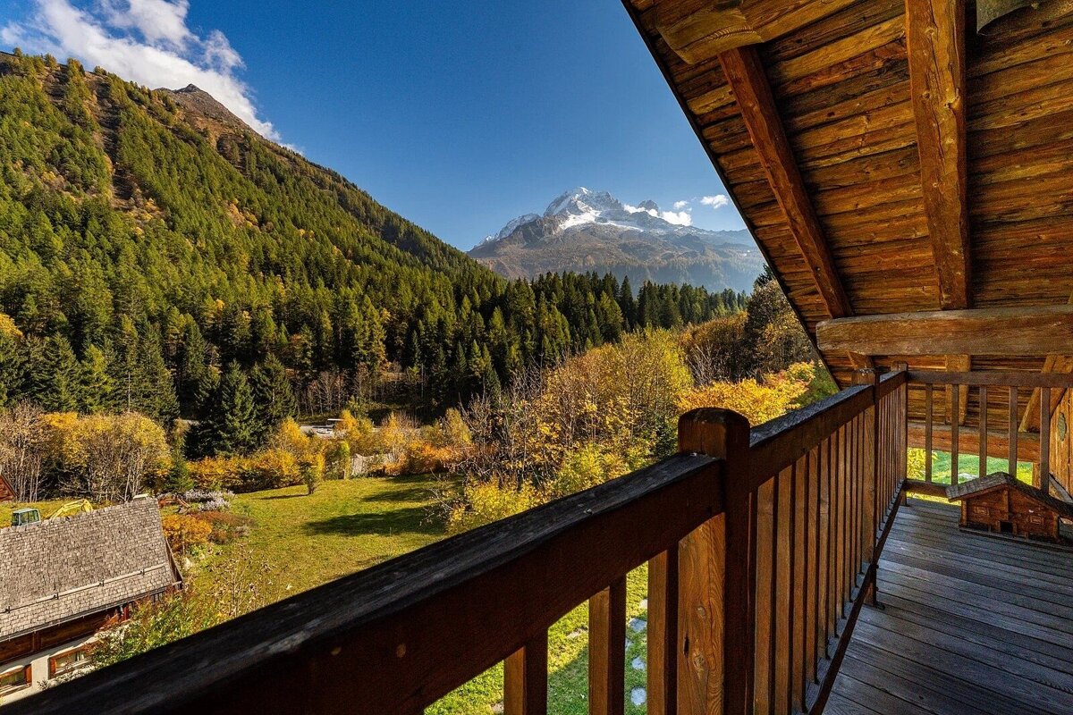 Cozy wooden balcony overlooking a vibrant autumn mountain valley with evergreen forests and a majestic snow-capped peak under a clear sky.
