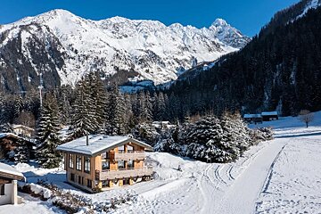 A wooden chalet nestled in a snowy alpine landscape with pine trees and majestic snow-capped mountains under a clear blue sky.