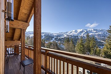 Wooden balcony overlooking majestic snow-capped mountains, a ski slope, and pine forests under a bright blue sky. Serene winter resort scene.