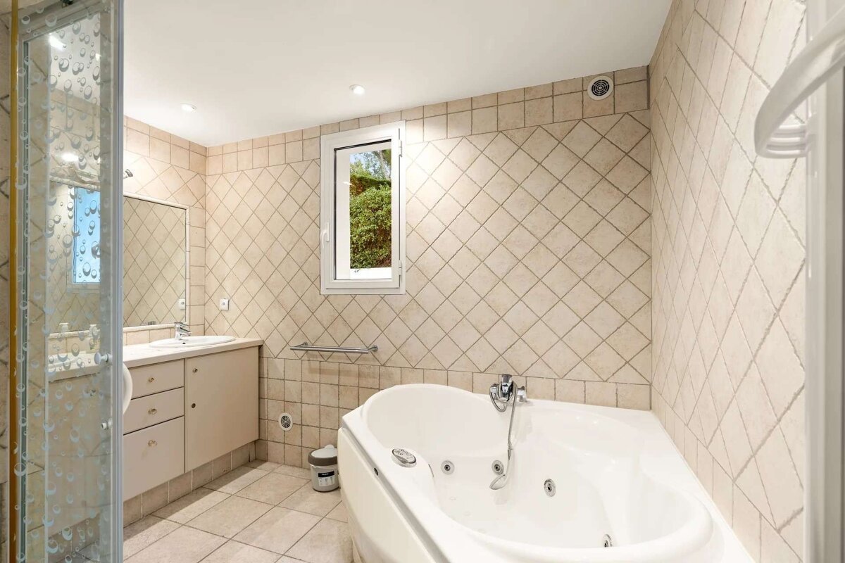 A tiled bathroom featuring a glass shower, vanity, corner jacuzzi tub, and a window looking out to greenery. Beige diagonal wall tiles.