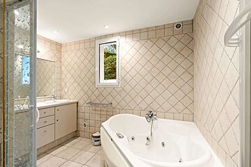 A tiled bathroom featuring a glass shower, vanity, corner jacuzzi tub, and a window looking out to greenery. Beige diagonal wall tiles.