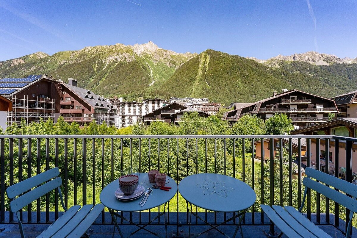A balcony with blue chairs and tables, offering a picturesque view of alpine chalets and green mountains under a bright blue sky.