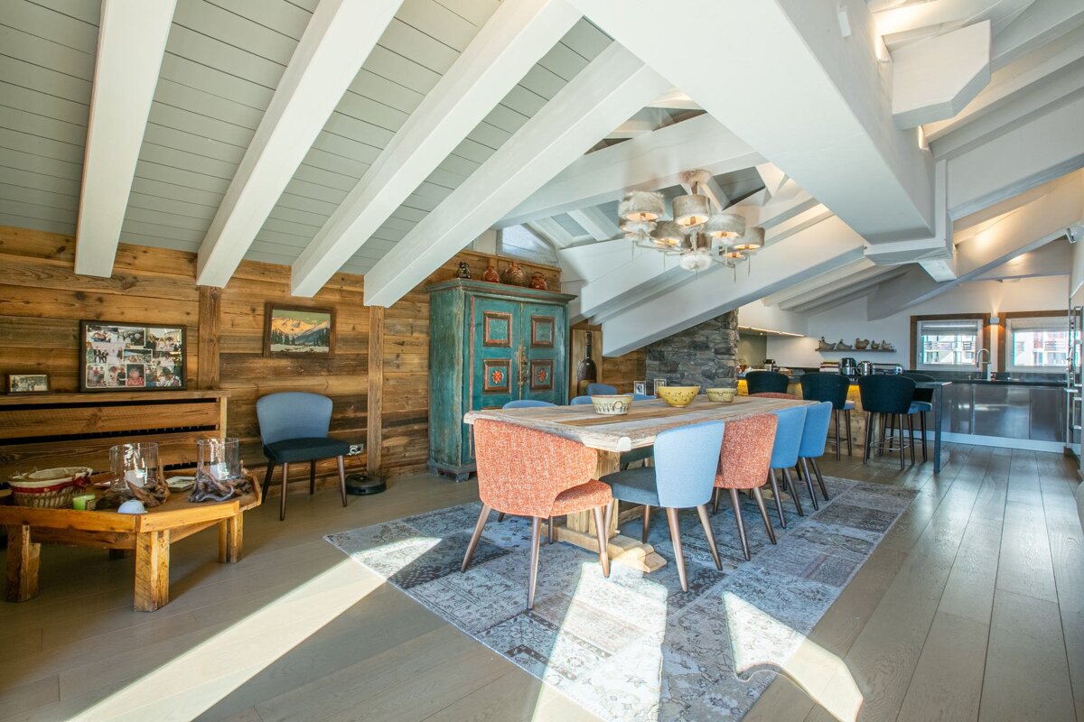 A rustic-modern chalet dining area with colorful chairs, a wooden table, wood-paneled walls, and white ceiling beams.