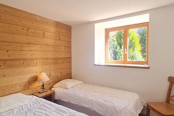 Cozy bedroom featuring two twin beds with white duvets, a rustic wood-paneled wall, and a window showing green trees outside.