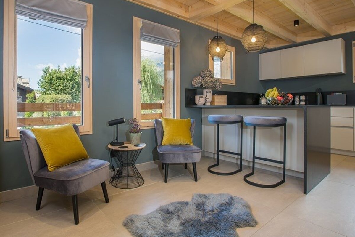 Cozy modern interior featuring grey walls, wooden ceilings, two grey armchairs with yellow cushions, and kitchen bar stools.