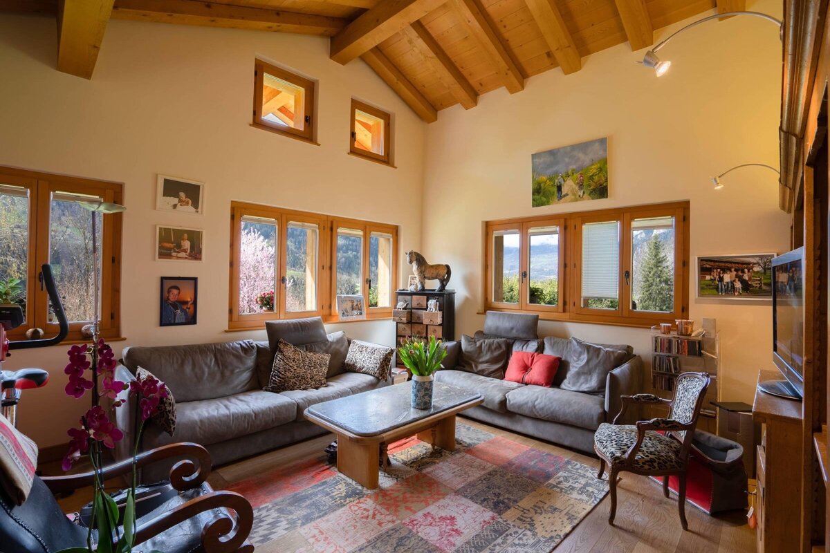 A cozy living room featuring grey sofas, a patterned rug, a wooden beamed ceiling, and large wooden-framed windows.