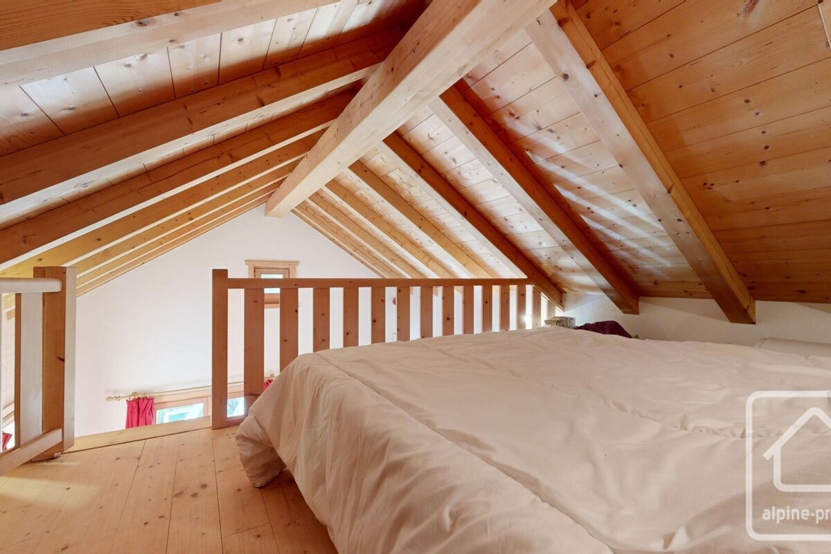 A cozy attic bedroom with exposed wooden beams, a white bed, and wooden floors, overlooking a lower level through a railing.