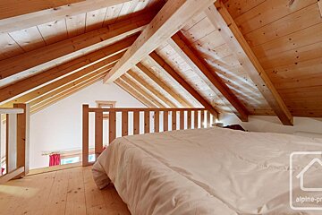 A cozy attic bedroom with exposed wooden beams, a white bed, and wooden floors, overlooking a lower level through a railing.