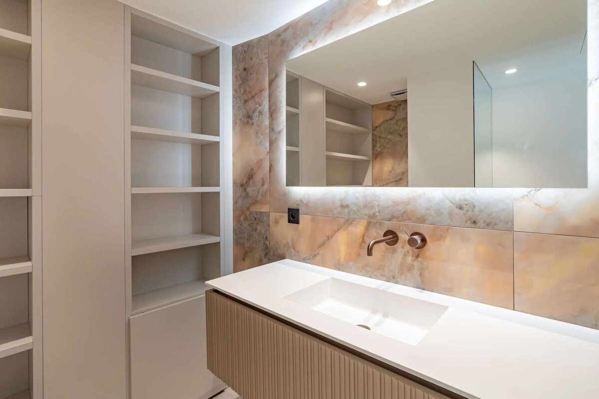 Stylish bathroom with white vanity & sink, copper faucet, backlit mirror, and marble wall tiles next to white shelving.