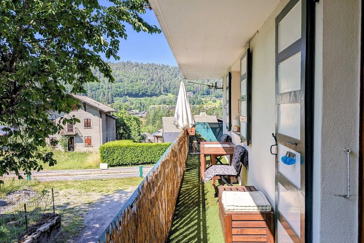 A narrow balcony with wooden furniture and artificial grass, overlooking a scenic green landscape with hills and houses.