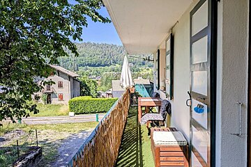 A narrow balcony with wooden furniture and artificial grass, overlooking a scenic green landscape with hills and houses.