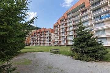 Two multi-story buildings with balconies, fronted by a green lawn, pine trees, and a bench, under a blue sky with distant mountains.