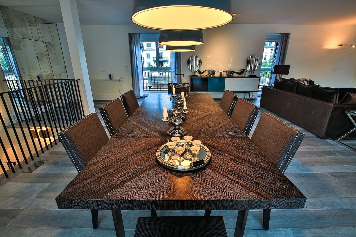An elegant, modern dining room features a long dark wooden table with upholstered chairs, under bright pendant lights. A living area and balcony are visible.