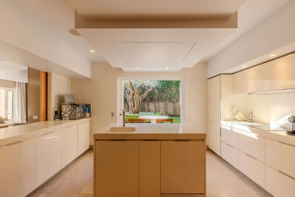 A modern, minimalist kitchen in cream tones with a central island and a large window looking out onto a lush green garden with a tree and table.