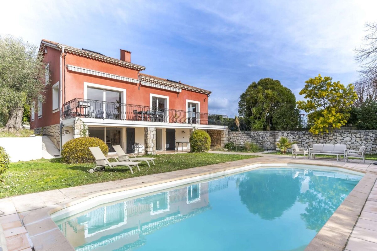 A striking red house with balconies overlooks a shimmering swimming pool and green lawn, bordered by a stone wall and trees under a clear blue sky.