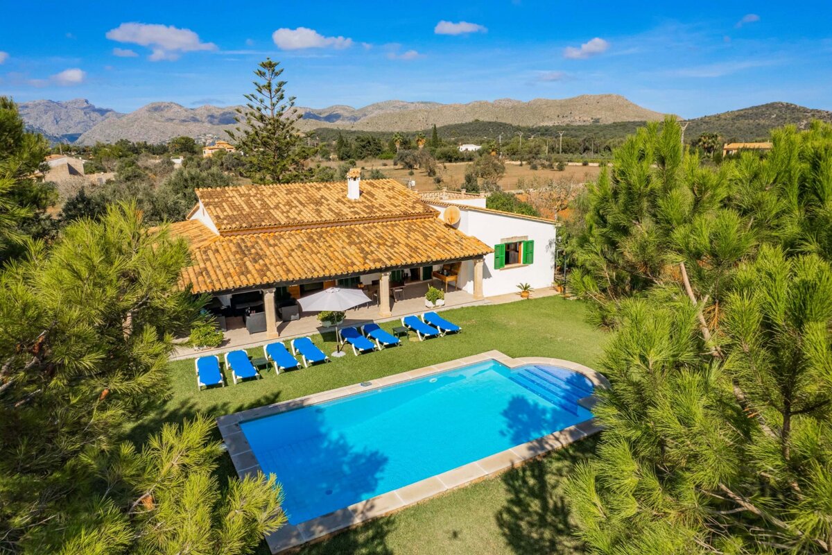 Sunny aerial view of a charming villa with a blue pool, sun loungers, and green lawn, surrounded by lush trees and distant mountains.