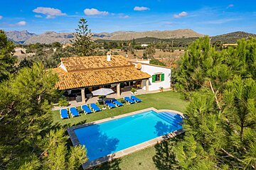 Sunny aerial view of a charming villa with a blue pool, sun loungers, and green lawn, surrounded by lush trees and distant mountains.