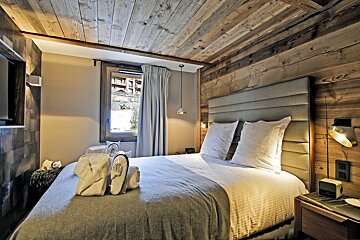 A cozy alpine bedroom featuring rustic wood-paneled walls and ceiling, a large bed with plush bedding, and a window with mountain views.
