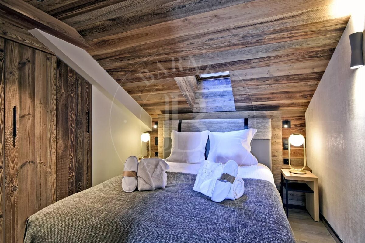 Cozy chalet bedroom with warm wooden walls and sloped ceiling, a bed with white linen, slippers, and towels, plus modern lighting.