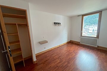 An empty room with a wooden floor, white walls, built-in closet, and a window overlooking a mountain town. It has a wall shelf and light fixture.