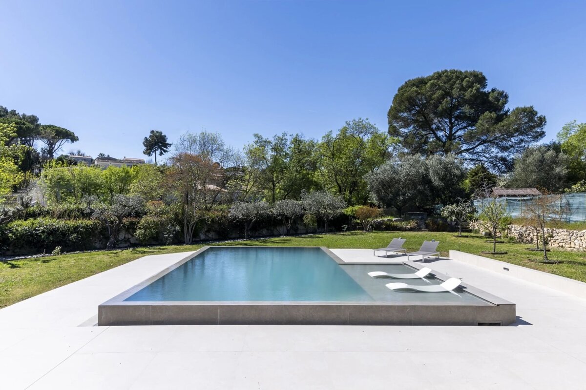 A modern rectangular pool with light decking, sun loungers, and green trees under a clear blue sky on a sunny day.