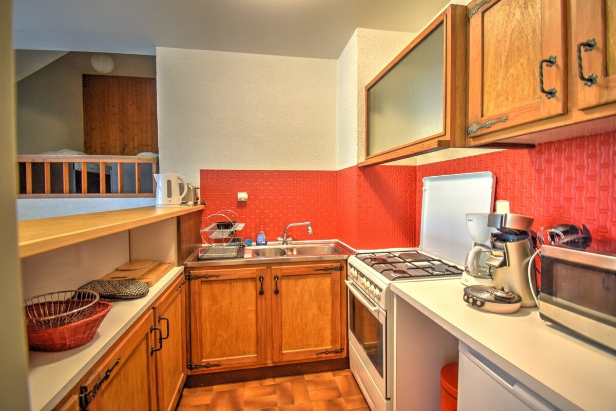 A small kitchen with red tiled backsplash, wooden cabinets, and various appliances. A loft bedroom with a wooden railing is visible in the background.