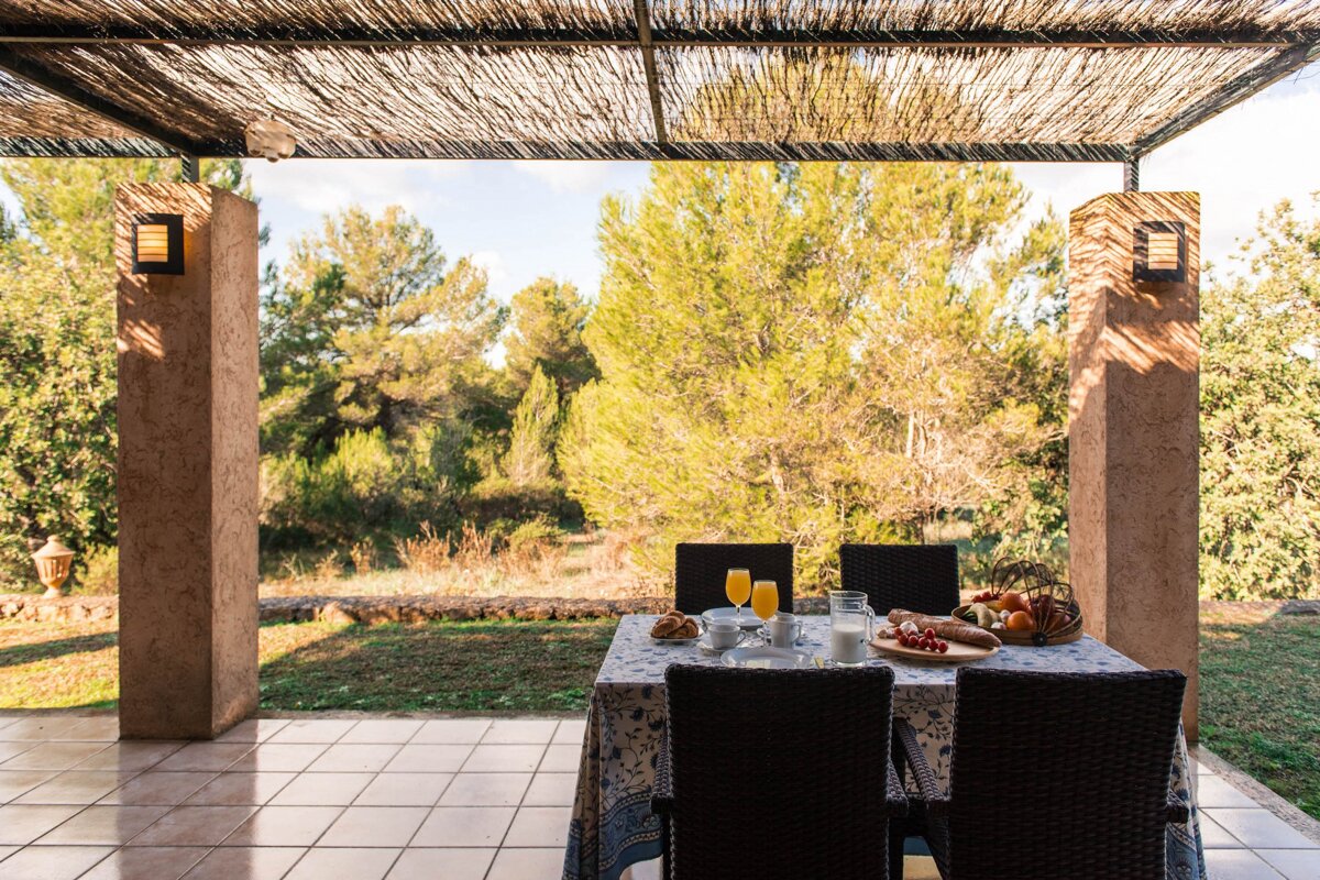 Breakfast for two on a shaded patio under a woven roof, overlooking a sunny forest. The table has juice, croissants, and fruit.