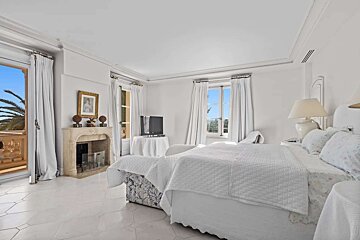 A bright, white bedroom with a large bed, fireplace, TV, and balcony access revealing palm trees and an outdoor view.