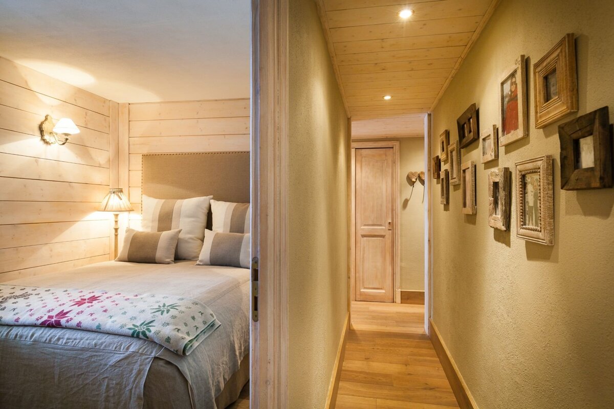 Cozy bedroom with wooden walls and bed seen through a doorway, next to a hallway with framed pictures and a wooden ceiling.