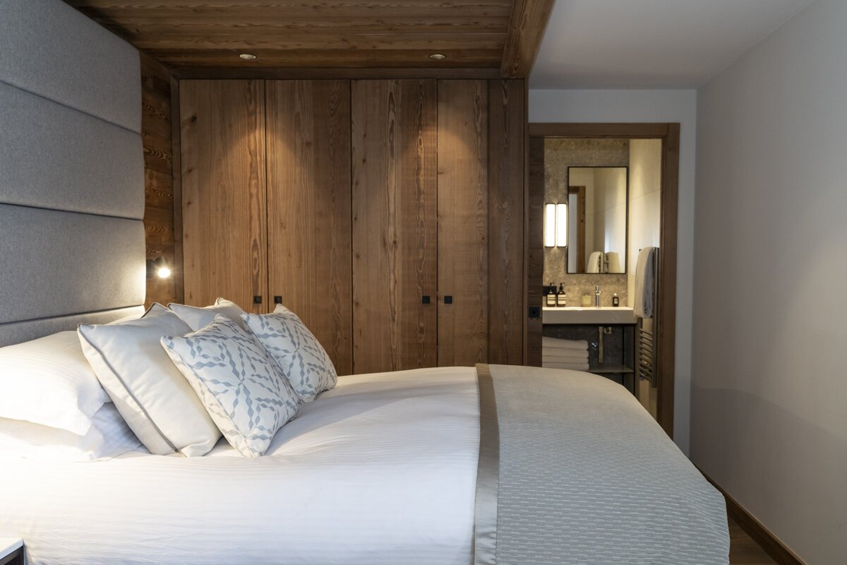 A cozy, modern bedroom featuring a plush white bed, patterned pillows, wooden built-in wardrobe, and an inviting glimpse into a chic bathroom.