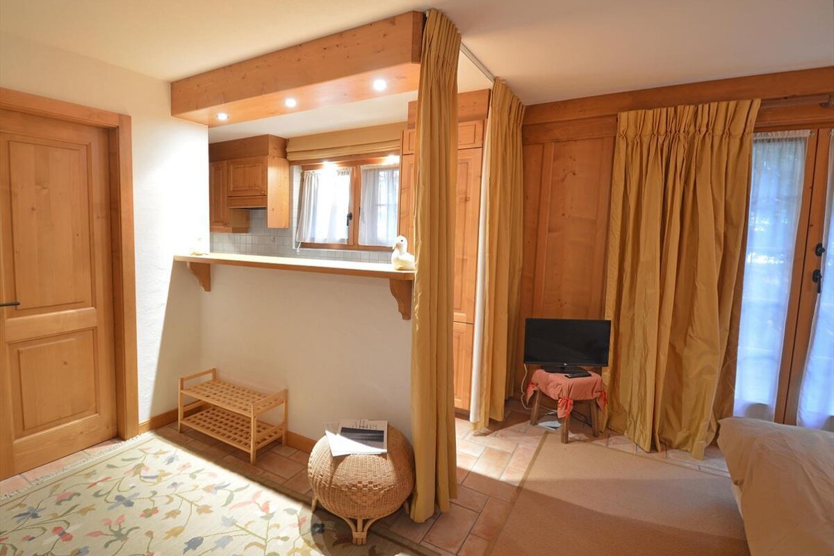 A cozy room with warm wood accents, a small kitchen counter, golden curtains, and a floral rug. A TV and wicker stool complete the inviting space.