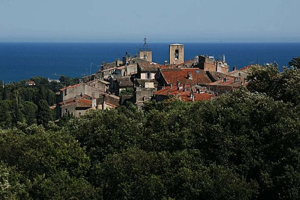 A picturesque village with red-tiled roofs and a bell tower emerges from green trees, overlooking the deep blue sea under a clear sky.