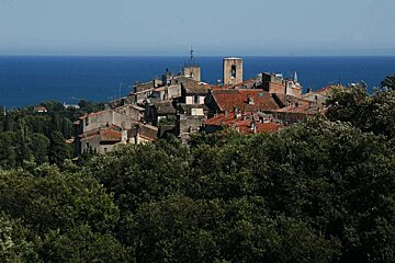 A picturesque village with red-tiled roofs and a bell tower emerges from green trees, overlooking the deep blue sea under a clear sky.