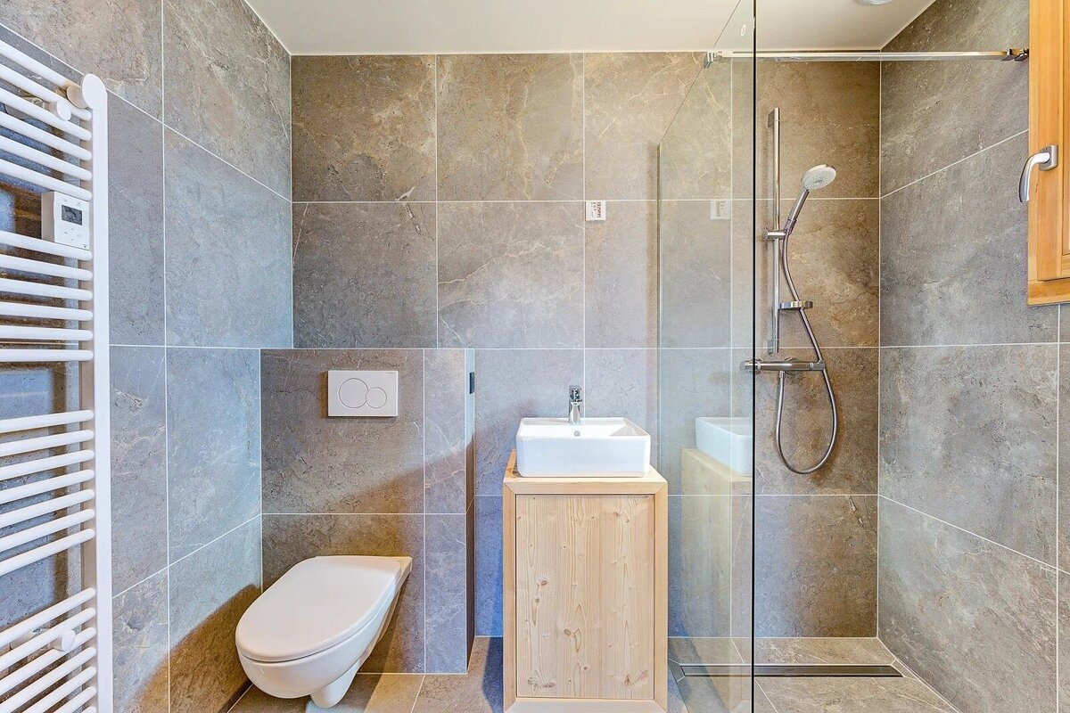 Modern bathroom with grey stone tiles, a wall-mounted toilet, a light wooden vanity, and a glass walk-in shower.