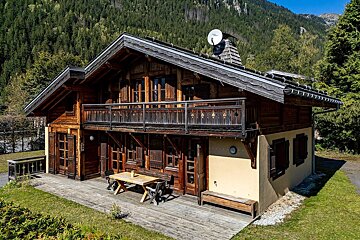 A rustic wooden chalet with a balcony, outdoor table, and satellite dish, set on a sunny day against a forested mountain backdrop.