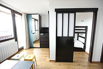 A modern room with wooden floors, large windows, colorful stools, and a sliding door to a bunk bed area. A dresser with a TV is visible.