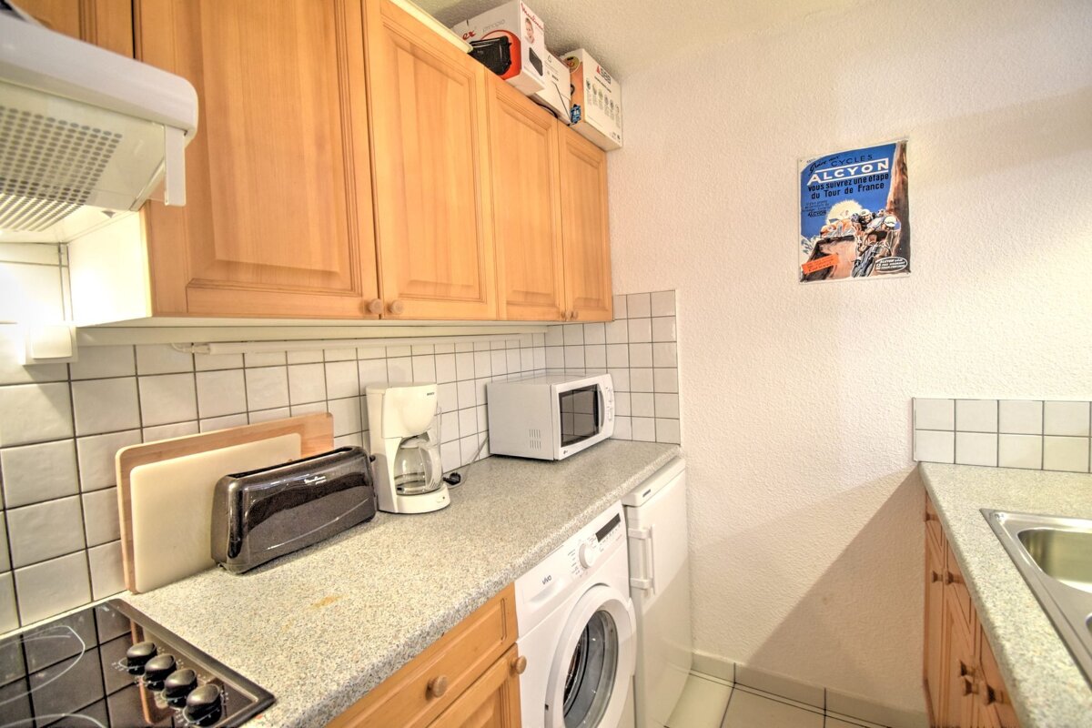 A functional kitchen with light wood cabinets, white tiled backsplash, and a counter holding a stove, toaster, coffee maker, microwave, and washing machine. A cycling poster is on the wall.
