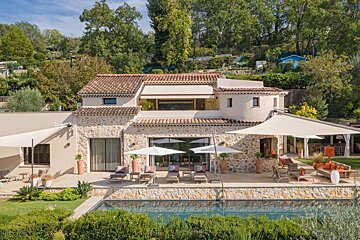 Luxurious stone villa with a terracotta roof, expansive patio, swimming pool, and sun loungers, set against a verdant hillside.