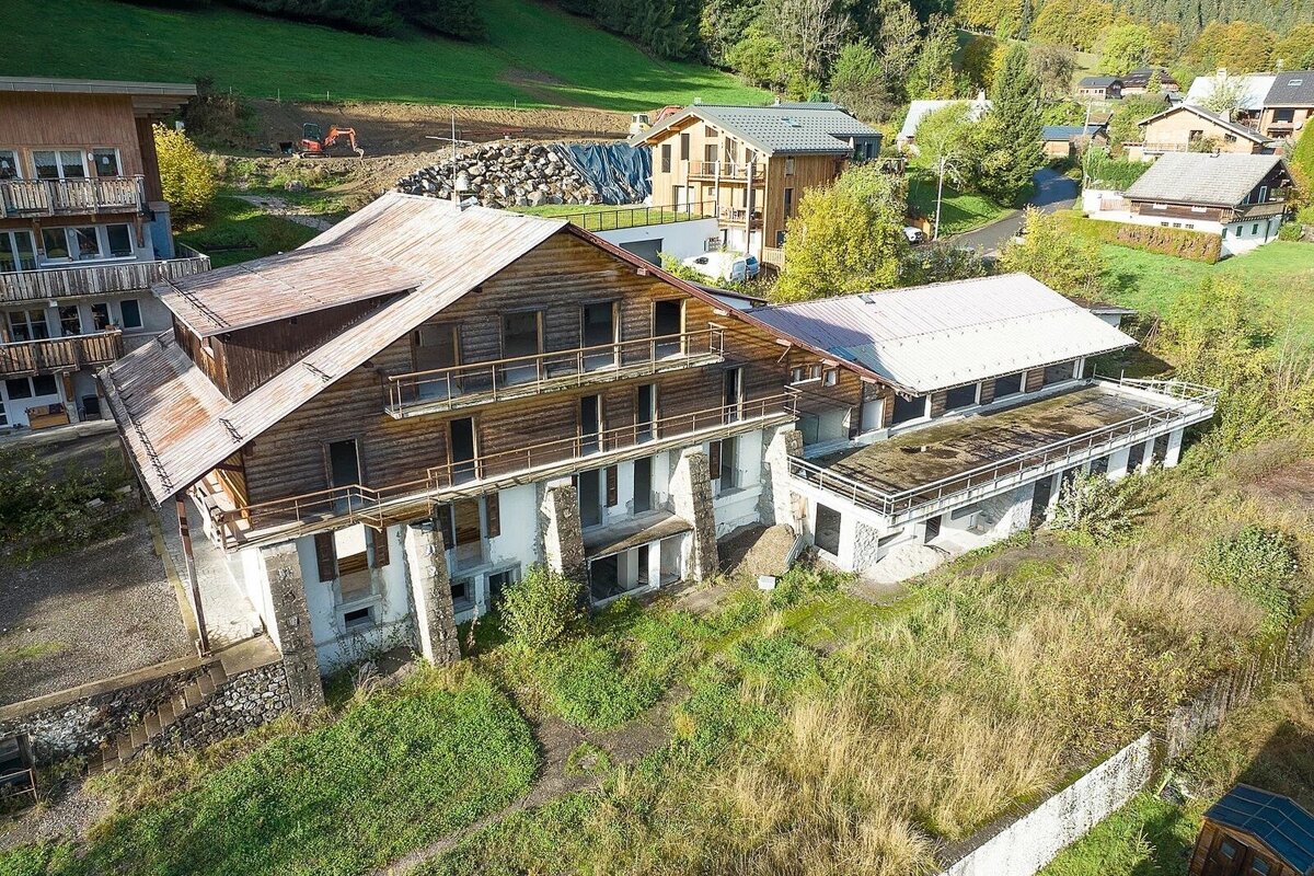 An aerial view shows a large, weathered multi-story building with wooden siding and stone foundations, appearing abandoned on a green hillside amidst other houses.