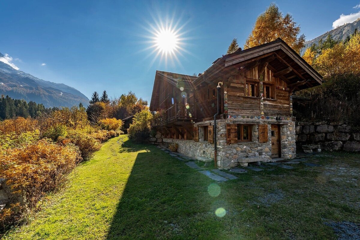 Rustic wooden chalet in a sunny autumn mountain landscape with vibrant yellow trees, green grass, and a clear blue sky.