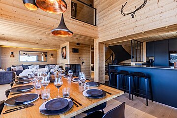 A modern wooden chalet dining area featuring a set table, copper pendant lights, and a sleek dark blue kitchen island.