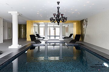 A luxurious indoor swimming pool featuring a black chandelier, white columns, golden mosaic wall, starry ceiling, and lounge chairs.